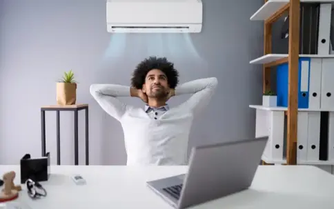 A man with curly hair relaxes under a wall-mounted air conditioner in an office, enjoying the cool air.
