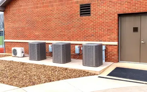 Three large HVAC units and a smaller mini-split unit are positioned against a red brick building, next to a brown door.
