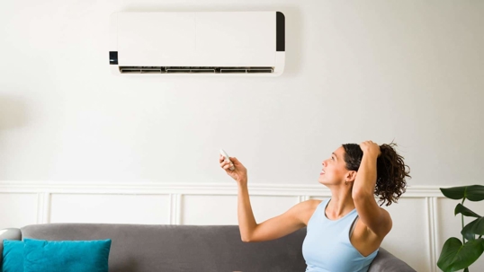 A woman with curly hair looks up at a white wall-mounted AC unit, holding a remote control.
