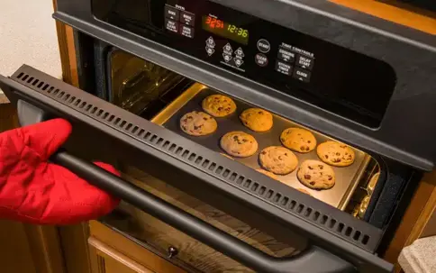 Oven mitt holds open an oven door, revealing a baking sheet with eight golden chocolate chip cookies