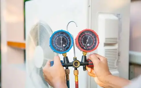 A technician's hands adjust a blue and a red manifold gauge set connected to an outdoor air conditioning unit for service.
