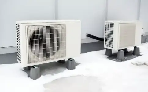 Two outdoor air conditioning units are shown in a snowy setting. They are light-colored, elevated on stands, and connected to a building wall.