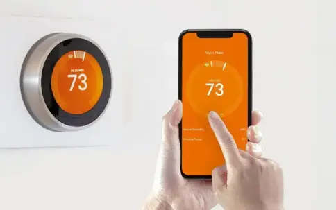 A hand uses a smartphone app to control a smart thermostat on a wall. Both the phone and thermostat display 73 degrees