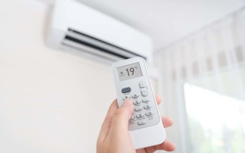 A hand holds an AC remote, showing 19°C, pointed towards a wall-mounted air conditioner.