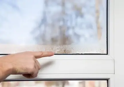 A hand points to severe condensation and ice forming on the inside of a window pane