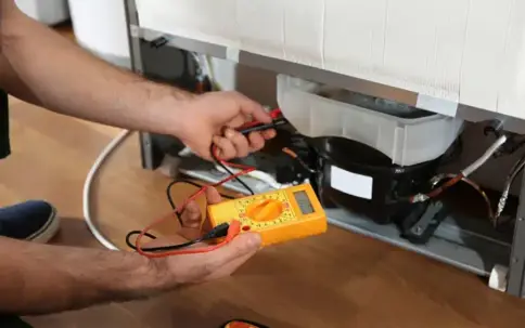 A person uses a yellow multimeter to test components at the back of a refrigerator.