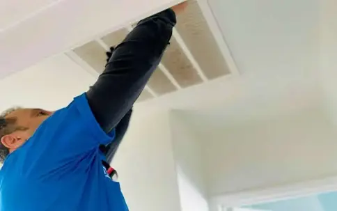 A technician in a blue shirt reaches up to service a ceiling air vent. This image demonstrates professional home maintenance.