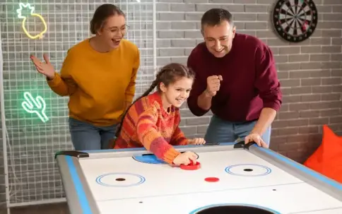 A happy family plays air hockey, with a child striking the puck as her parents cheer excitedly beside her.
