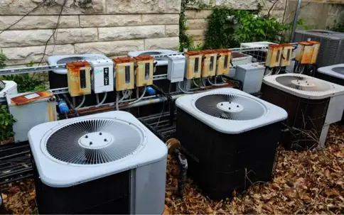 Multiple outdoor air conditioning units are shown with a stone wall backdrop and dried leaves.