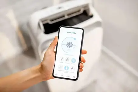 A hand holds a smartphone displaying a ventilation app controlling a white smart air device.