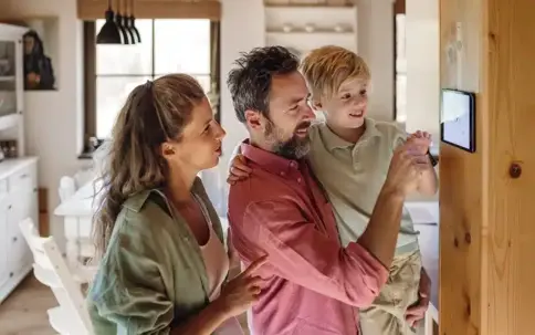 A happy family, a man, woman, and child, interact with a wall-mounted smart home display.