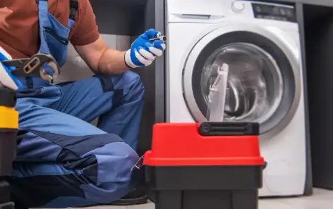 A person in blue overalls repairs a washing machine, holding tools next to a red toolbox.