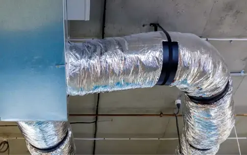 HVAC ducts, showing a rectangular metal duct connecting to a curving, insulated flexible silver duct, mounted against a concrete ceiling.