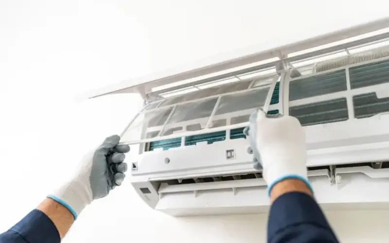 A close-up shows hands using a screwdriver to repair an open, white, wall-mounted air conditioner. This image illustrates AC repair or its associated costs.