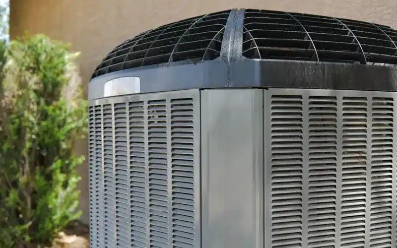 A home's outdoor air conditioning unit, light grey with louvered sides and a dark top, stands next to a green bush.