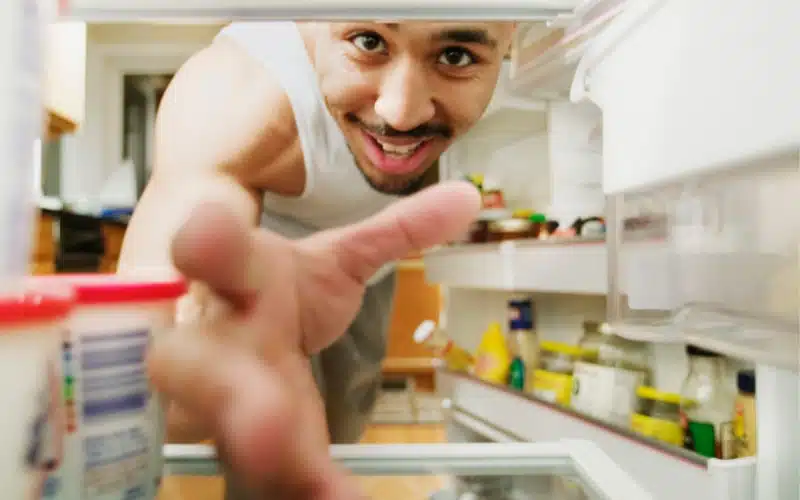 A smiling man, viewed from inside a refrigerator, reaches out towards the camera.