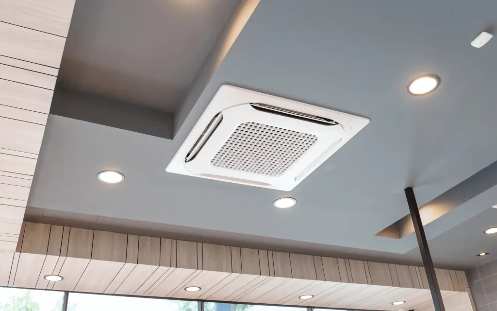 A white ceiling cassette air conditioner unit is mounted centrally on a gray ceiling, surrounded by recessed lighting and modern wooden paneling.