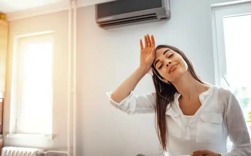 A woman wipes her forehead, looking uncomfortable from the heat in a room with an air conditioner.