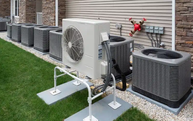 A row of various outdoor HVAC condenser units, including a large white unit on a stand, showcasing different system sizes next to a building. 