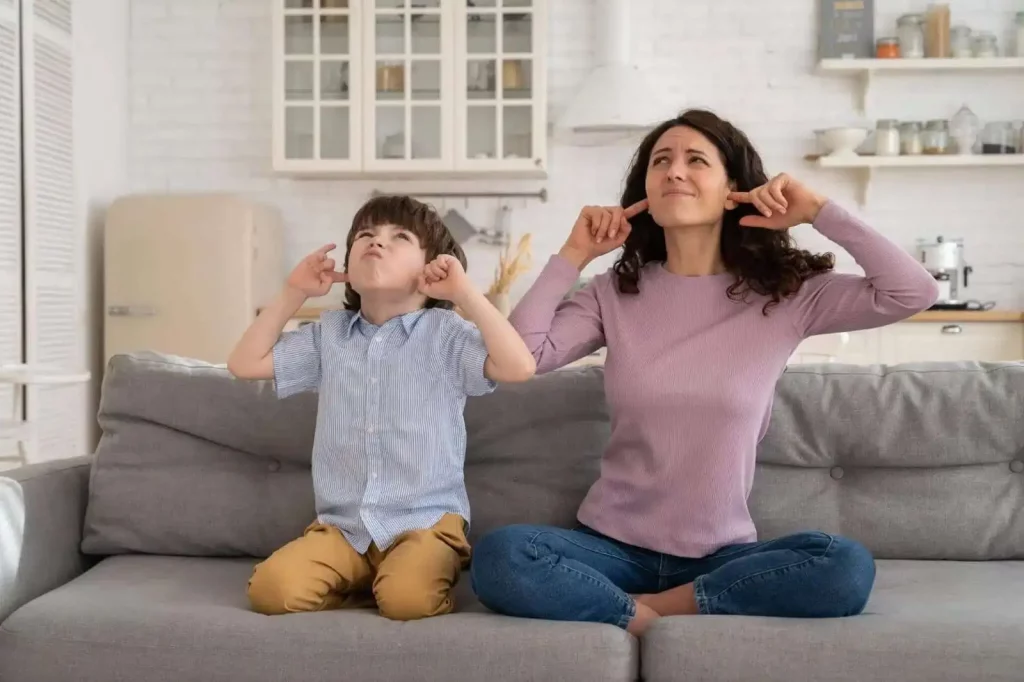 A mother and son on a couch cover their ears with exasperated expressions, illustrating discomfort from a loud noise, such as a noisy furnace.