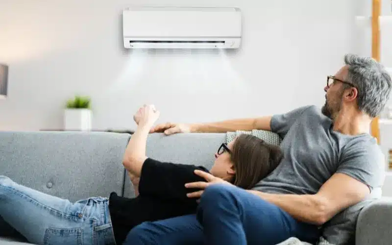 A couple relaxes comfortably on a couch with a white wall-mounted air conditioner cooling the room
