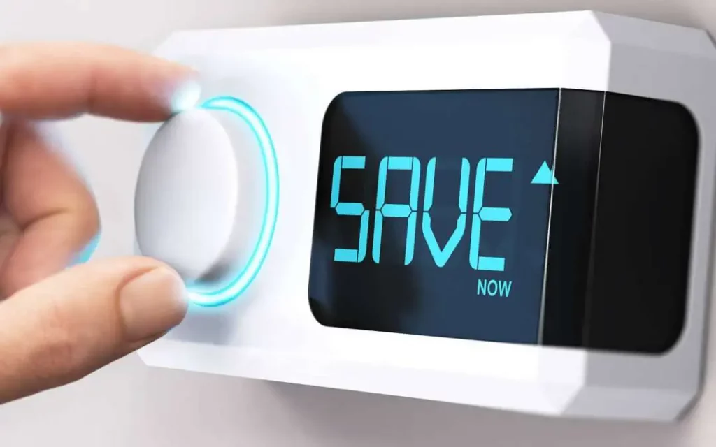 A hand turns a modern smart thermostat knob, featuring a blue glowing ring, with "SAVE NOW" displayed on its screen, suggesting energy savings.