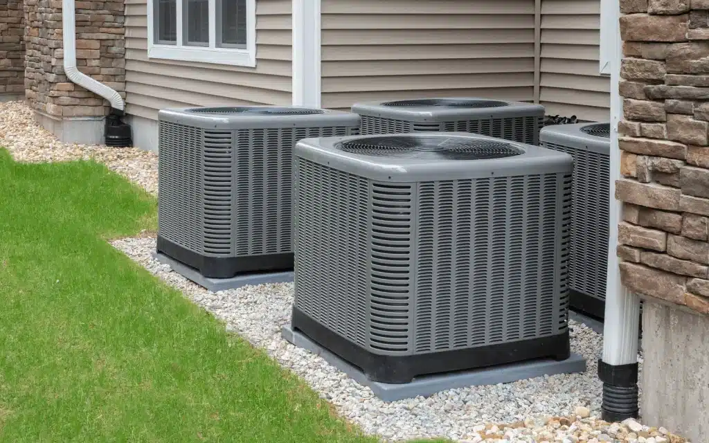 Four gray outdoor air conditioning units are neatly arranged on a gravel bed beside a house.