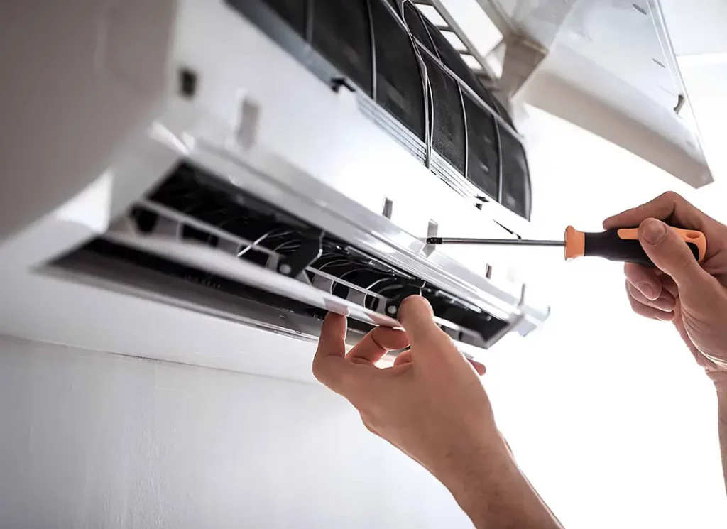 A close-up shows hands using a screwdriver to repair an open, white, wall-mounted air conditioner. This image illustrates AC repair or its associated costs.